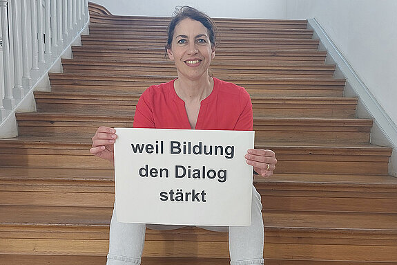 Stefanie Schwegler feiert die Demokratie, weil Bildung den Dialog stärkt. Foto: Schwegler Eine Frau mit dunklen Haaren und rotem Oberteil sitzt auf einer Holztreppe und hält ein Schild.