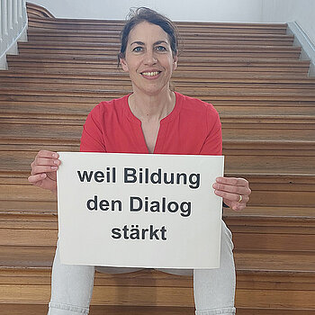 Stefanie Schwegler feiert die Demokratie, weil Bildung den Dialog stärkt. Foto: Schwegler Eine Frau mit dunklen Haaren und rotem Oberteil sitzt auf einer Holztreppe und hält ein Schild.