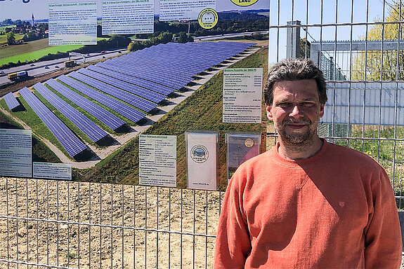 Ein Mann im orangerotem Pullover vor einem Zaum mit einem Schild der Freiflächen-Solaranlage "Bürger-Solarpark-Johanneck"