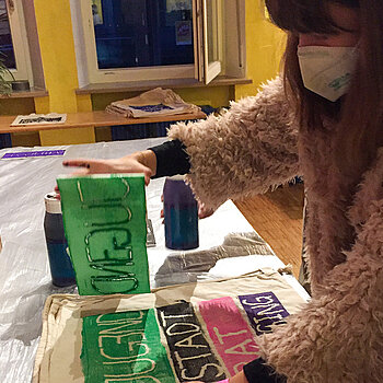 Junge Frau mit Maske zieht eine grüne Druckvorlage von einer Stofftasche ab.