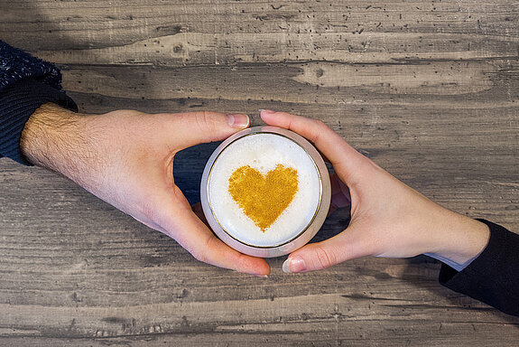 Man  sieht, wie eine Männerhand und eine Frauenhand ein Glas mit Milchschaum zwischen sich halten. Auf dem Milchschaum befindet sich ein goldenes Herz.