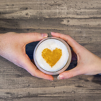 Man  sieht, wie eine Männerhand und eine Frauenhand ein Glas mit Milchschaum zwischen sich halten. Auf dem Milchschaum befindet sich ein goldenes Herz.