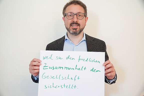 Stadtratsmitglied Simon Schindlmayr. Foto: Stadt Freising Ein Mann mit Brille und einem blauen Hemd und einem Jackett drüber zeigt ein Schild mit der Aufschrift "weil sie den friedlichen Zusammenhalt der Gesellschaft sicherstellt."