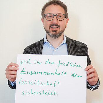 Stadtratsmitglied Simon Schindlmayr. Foto: Stadt Freising Ein Mann mit Brille und einem blauen Hemd und einem Jackett drüber zeigt ein Schild mit der Aufschrift "weil sie den friedlichen Zusammenhalt der Gesellschaft sicherstellt."