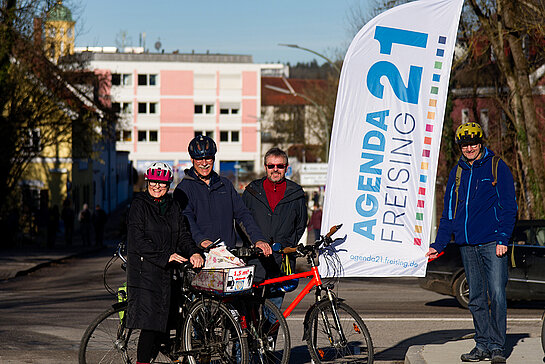 4 Menschen mit Rädern und teiweise Radhelmen sowie einer Beach-Flag "Agenda21" stehen auf der Brücke. Im Hintergrund ein rosa Haus