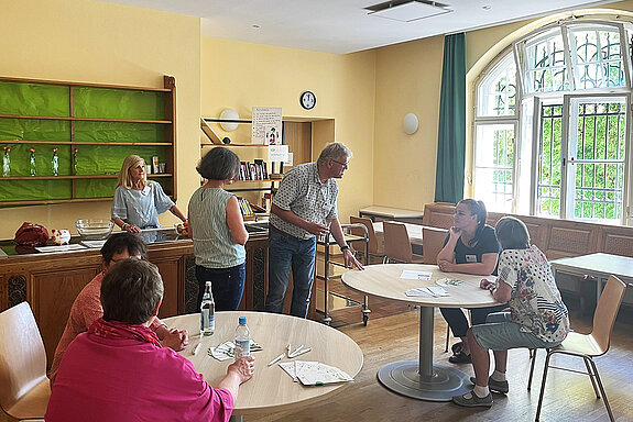 hinter der Theke steht eine Frau, davor stehen eine Frau und ein Mann, die sich mit zwei Personen an einem Tisch zuwenden. Weiter vorne noch ein Tisch mit 2 Personen im Gespräch.