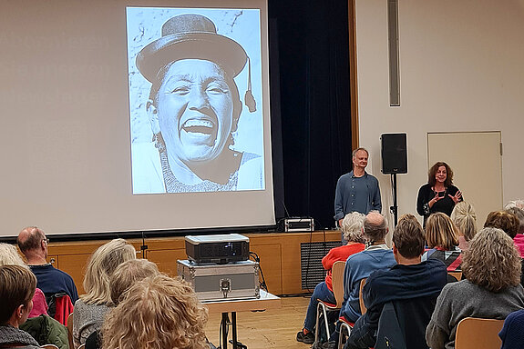 Ein Bild einer lachenden Indio-Frau auf der Leinwand vor sitzendem Publikum von hinten. Eine Frau und ein Mann sprechen zum Publikum.