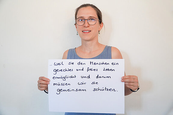 Stadtratsmitglied Ricarda Schindler. Foto: Stadt Freising Eine Frau mit Brille in einem blauen tank Top zeigt ein Schild mit der Aufschrift "weil sie den Menschen ein gerechtes und freies Leben ermöglicht und darum müssen wir sie gemeinsam schützen."