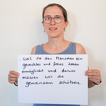 Stadtratsmitglied Ricarda Schindler. Foto: Stadt Freising Eine Frau mit Brille in einem blauen tank Top zeigt ein Schild mit der Aufschrift "weil sie den Menschen ein gerechtes und freies Leben ermöglicht und darum müssen wir sie gemeinsam schützen."