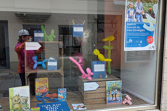 Kisten und blaue Papiertüten, in denen Spielideen versteckt sind. Dazu ein Plakat und Ballonfiguren.