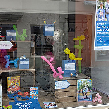 Kisten und blaue Papiertüten, in denen Spielideen versteckt sind. Dazu ein Plakat und Ballonfiguren.