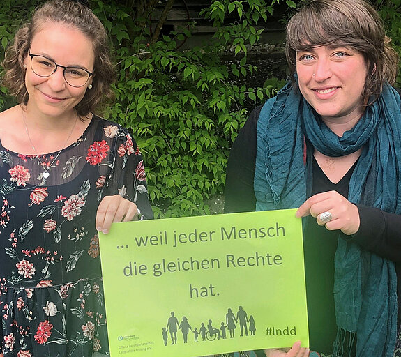 Alexandra Deml und Saskia Hobmeier halten ein grünes Plakat mit der Aufschrift "... weil jeder Mensch die gleichen Rechte hat." Dazu sieht man Silhouetten von verschiedensten Menschen. 