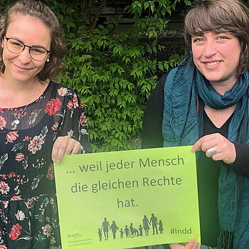 Alexandra Deml und Saskia Hobmeier halten ein grünes Plakat mit der Aufschrift "... weil jeder Mensch die gleichen Rechte hat." Dazu sieht man Silhouetten von verschiedensten Menschen. 