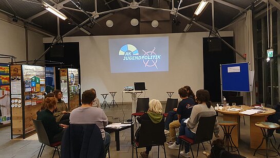 Eine gruppe junger Menschen sitzt im Kreis. Im Hintergrund ist das Kreisjugendring-Logo an die Wand projiziert. Daneben die Module der Ausstellung