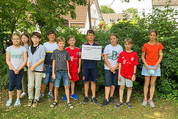 Schülerinnen und Schüler stehen vor einer Hecke. Ein Schüler hält das Schild mit der Aufschrift: "... jeder Mensch gleich viel zählt"