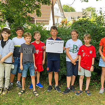 Schülerinnen und Schüler stehen vor einer Hecke. Ein Schüler hält das Schild mit der Aufschrift: "... jeder Mensch gleich viel zählt"