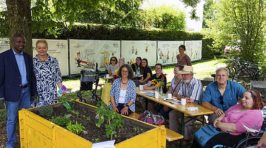 Eine Gruppe Menschen an Biertischen unter Bäumen, im Hintergrund Bauzaunbanner mit Cartoons, im Vordergrund ein Hochbeet.
