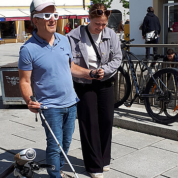 Eine Frau hilft einen Mann mit Blindensimulationsbrille und Blindenstock