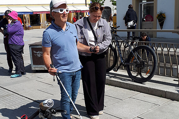 Eine Frau hilft einen Mann mit Blindensimulationsbrille und Blindenstock