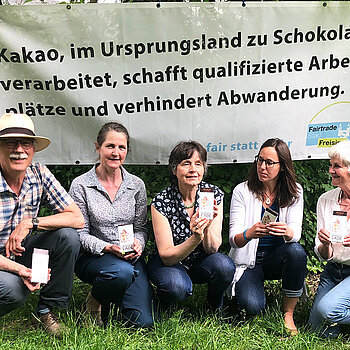 vier Frauen und ein Mann hocken vor einem Banner mit dem Text: Kakao, im Ursprungsland zu Schokolade verarbeitet, schafft qualifizierte Arbeitsplätze und verhindert Abwanderung.