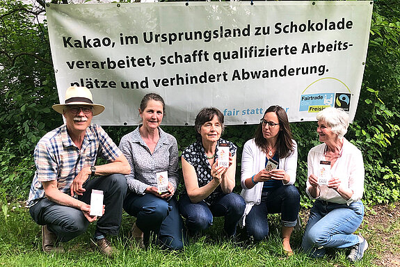vier Frauen und ein Mann hocken vor einem Banner mit dem Text: Kakao, im Ursprungsland zu Schokolade verarbeitet, schafft qualifizierte Arbeitsplätze und verhindert Abwanderung.