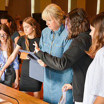 Eine Frau hält ein Tablet in der Hand, Schülerinnen neben ihr zeigen, wie es funktioniert.
