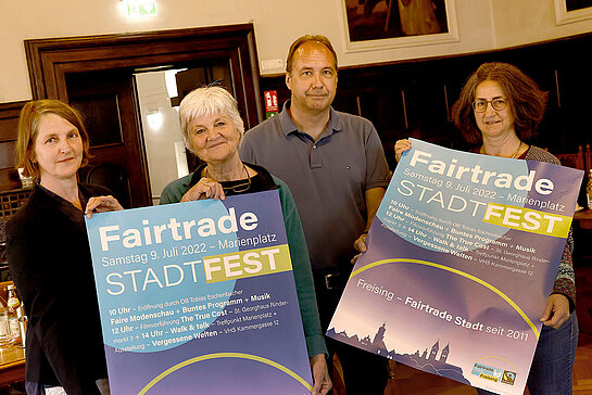 drei Frauen und ein Mann halten in einem dunkel getäfelten Raum zwei große Plakate "Fairtrade-Stadtfest"