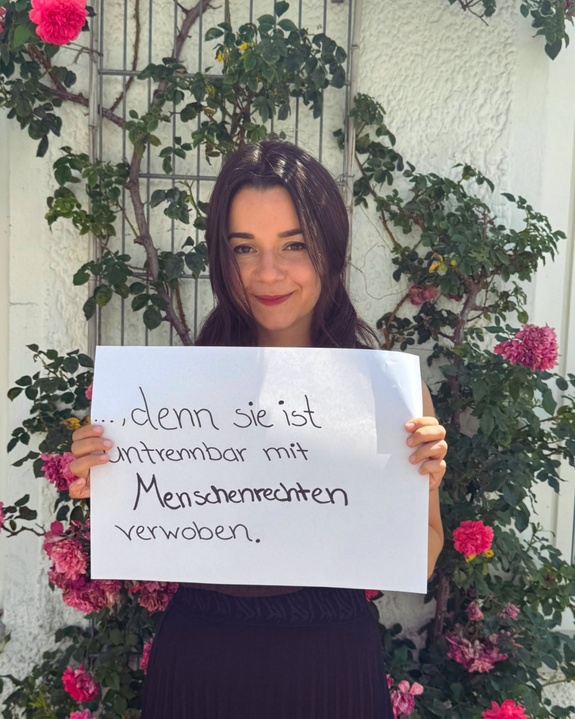 Mirjam Mayer (Kreisjungendring Freising) feiert die Demokratie, denn sie ist untrennbar mit Menschenrechten verwoben. Foto: KJR Freising Eine Frau mit dunklen langen Haaren steht vor einer Wand mit einer Kletterrose und hält ein Schild.