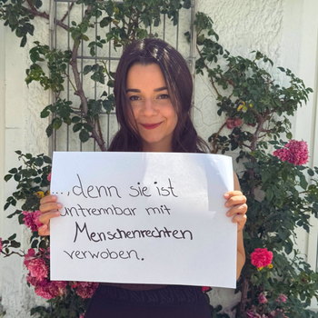 Mirjam Mayer (Kreisjungendring Freising) feiert die Demokratie, denn sie ist untrennbar mit Menschenrechten verwoben. Foto: KJR Freising Eine Frau mit dunklen langen Haaren steht vor einer Wand mit einer Kletterrose und hält ein Schild.