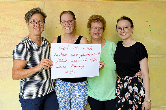 A. Kessel, L. Deuter, S. Böck und F. Attenkofer vom OMG Neufahrn feiern die Demokratie, weil sie sich sicher und geschützt fühlen, wenn sie offen ihre Meinung sagen. Foto: D. Lepkova 4 Frauen unterschiedlichen Alters stehen lächelnd vor einer gelben Wand und halten gemeinsam ein Schild hoch.