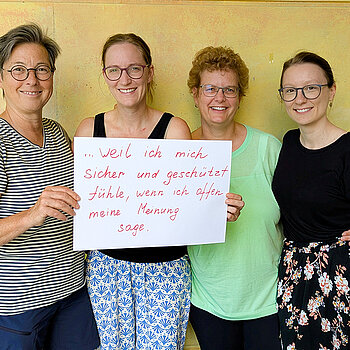 A. Kessel, L. Deuter, S. Böck und F. Attenkofer vom OMG Neufahrn feiern die Demokratie, weil sie sich sicher und geschützt fühlen, wenn sie offen ihre Meinung sagen. Foto: D. Lepkova 4 Frauen unterschiedlichen Alters stehen lächelnd vor einer gelben Wand und halten gemeinsam ein Schild hoch.