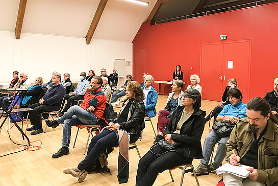 Menschen sitzen auf Stühlen in einem Saal.