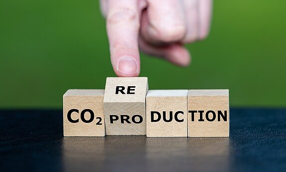 Vier Holzwürfel mit der Aufschrift CO2-PRO-DUC-TION vor einem grünen Hintergrund. Eine Hand stupst den zweiten Würfel von "PRO" auf die nächste Seite "RE" - so entsteht das Wort "CO2-Reduktion"