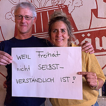 Franzi Kreuther und Ludwig Dinzinger (Furtner-Wirte) feiern die Demokratie, weil Freiheit nicht selbstverständlich ist. Foto: Claudia Bauer Ein Mann mit Brille in dunklem Shirt und eine Frau im gelben Pullover stehtn vor einem großen roten Gasthof-Logo und halten ein Schild.