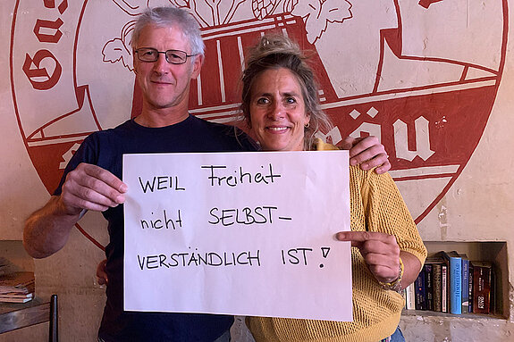 Franzi Kreuther und Ludwig Dinzinger (Furtner-Wirte) feiern die Demokratie, weil Freiheit nicht selbstverständlich ist. Foto: Claudia Bauer Ein Mann mit Brille in dunklem Shirt und eine Frau im gelben Pullover stehtn vor einem großen roten Gasthof-Logo und halten ein Schild.