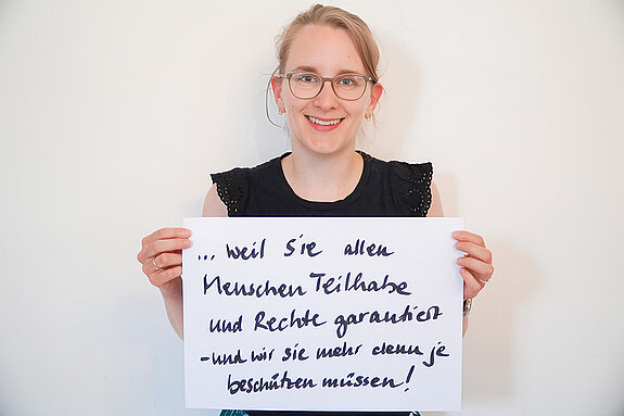Stadtratsmitglied Teresa Degelmann. Foto: Stadt Freising Eine Frau mit Brille und einem schwarzen T-Shirt zeigt ein Schild mit der Aufschrift "weil sie allen Menschen Teilhabe und Rechte gewährt- und wir sie mehr denn je beschützen müssen!"