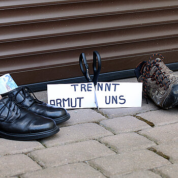 Links schicke schwarze Schuhe mit Scheinen darin, rechts schäbige Turnschuhe. Dazwischen ein Schild mit der Aufschrift." Armut trennt uns" das von einer Schere durchschnitten wird.