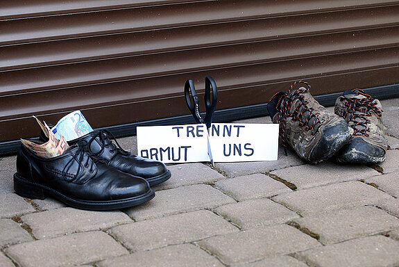 Links schicke schwarze Schuhe mit Scheinen darin, rechts schäbige Turnschuhe. Dazwischen ein Schild mit der Aufschrift." Armut trennt uns" das von einer Schere durchschnitten wird.