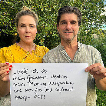 Carolin Nünemann (Pressesprecherin Stadt Freising und PR-Agentin) & Jens Nünemann (Schauspieler, Handwerker und Business-Coach) eine Frau mit dunkelblonden, hochgesteckten Haaren und ein Mann mit dunklen kurzen Haaren stehen vor einem Apfelbaum und halten ein Schild