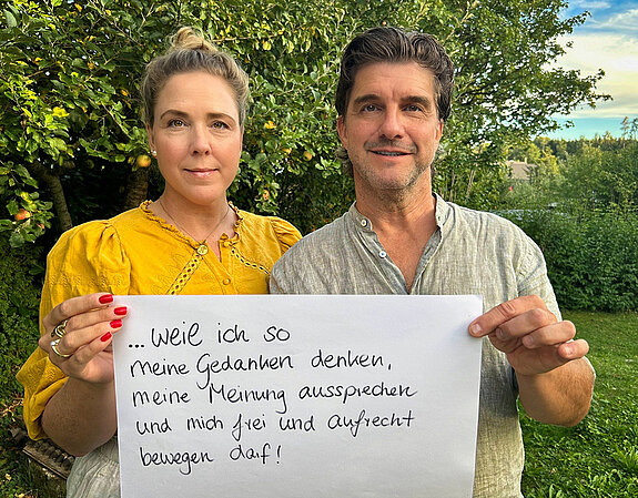 Carolin Nünemann (Pressesprecherin Stadt Freising und PR-Agentin) & Jens Nünemann (Schauspieler, Handwerker und Business-Coach) eine Frau mit dunkelblonden, hochgesteckten Haaren und ein Mann mit dunklen kurzen Haaren stehen vor einem Apfelbaum und halten ein Schild