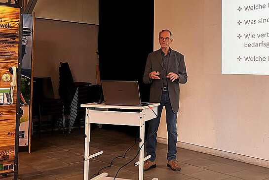 Ein Mann steht mit halb erhobenen Händen zwischen einer Präsentation und einem Ausstellungsmodul. Vor sich ein Tisch mit Laptop.