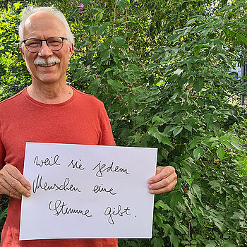 Jürgen Maguhn (vhs Freising und Agenda21) feiert die Demokratie, weil sie jedem eine Stimme gibt. Foto: Jürgen Maguhn Ein grauhaariger Mann mit Schnurbart und rotem T-Shirt steht vor einem Busch und hält ein Schild hoch.