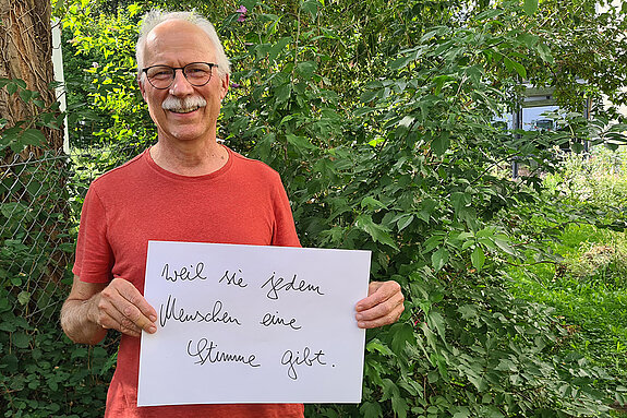 Jürgen Maguhn (vhs Freising und Agenda21) feiert die Demokratie, weil sie jedem eine Stimme gibt. Foto: Jürgen Maguhn Ein grauhaariger Mann mit Schnurbart und rotem T-Shirt steht vor einem Busch und hält ein Schild hoch.