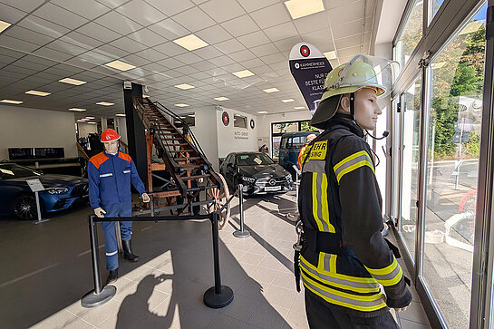 Die Freiwillige Feuerwehr bestückte das Fenster bei Kammermeier mit "Feuerwehrleuten" und Gerätschaften. Foto: Stadt Freising Zwei Schaufensterpuppen in Feuerwehr-Montur, eine historische Drehleiter und mehrere Fahrzeuge im Hintergrund.