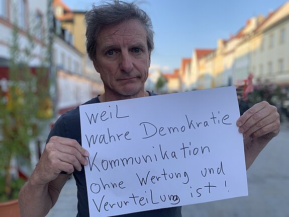 Norbert Bürger (Musiker) feiert die Demokratie, weil wahre Demokratie Kommunikation ohne Wertung und Verurteilung ist. Foto: Claudia Bauer Ein Mann mit kurzen dunkelblonden Haaren steht in der Straße und hält ein Schild