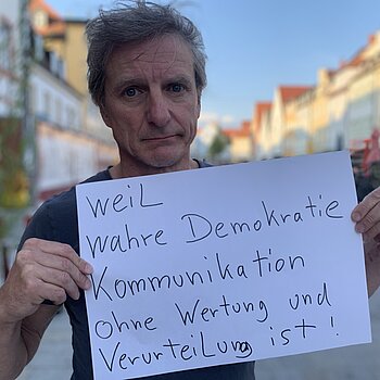 Norbert Bürger (Musiker) feiert die Demokratie, weil wahre Demokratie Kommunikation ohne Wertung und Verurteilung ist. Foto: Claudia Bauer Ein Mann mit kurzen dunkelblonden Haaren steht in der Straße und hält ein Schild