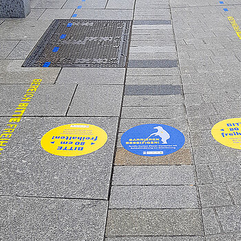 gelbe und blaue Markierungen auf dem grauen Pflaster, die den Leitstreifen für Sehbehinderte hervorheben. 