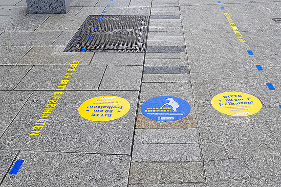 gelbe und blaue Markierungen auf dem grauen Pflaster, die den Leitstreifen für Sehbehinderte hervorheben. 