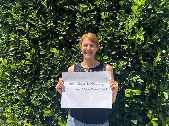 Birgit Irrgang (Kreisjugendring) feiert die Demokratie, weil Freiheit und Mitbestimmung keine Selbstverständlichkeit sind. Foto: kjr Eine dunkelblonde Frau im blauen Shirt steht vor einer Hecke und hält ein Schild.