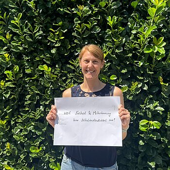 Birgit Irrgang (Kreisjugendring) feiert die Demokratie, weil Freiheit und Mitbestimmung keine Selbstverständlichkeit sind. Foto: kjr Eine dunkelblonde Frau im blauen Shirt steht vor einer Hecke und hält ein Schild.